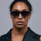 Brutal 2.0 Sunglasses by Tribu Eyewear — Grey grad polarised lens in a black round acetate frame on female grey background.