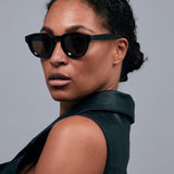 Brutal 4.0 Polarised Sunglasses by Tribu Eyewear — Black Round acetate frame, brown lens, on female light grey background.