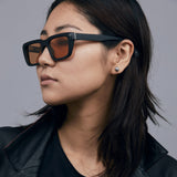 Brutal 5.0 Sunglasses by Tribu Eyewear — Matte black rectangular acetate frame, desert orange lens, on female light grey background.